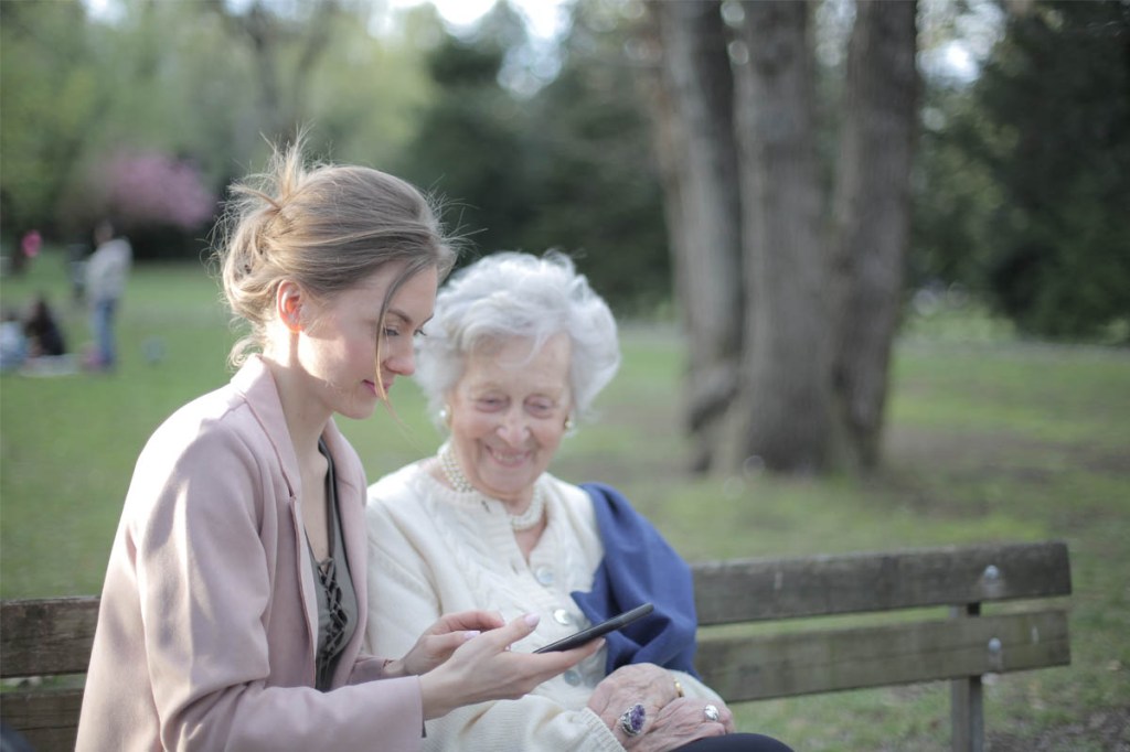 Eine jüngere und eine ältere Fraue sitzen zusammen im Park und schauen auf ein Smartphone.
