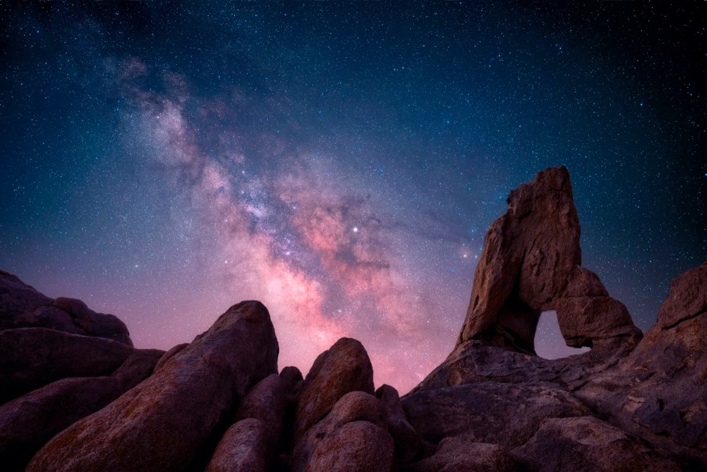 Ein Foto nach oben gerichtet zwischen Felsen auf dem der Sternenhimmel in pink-blau