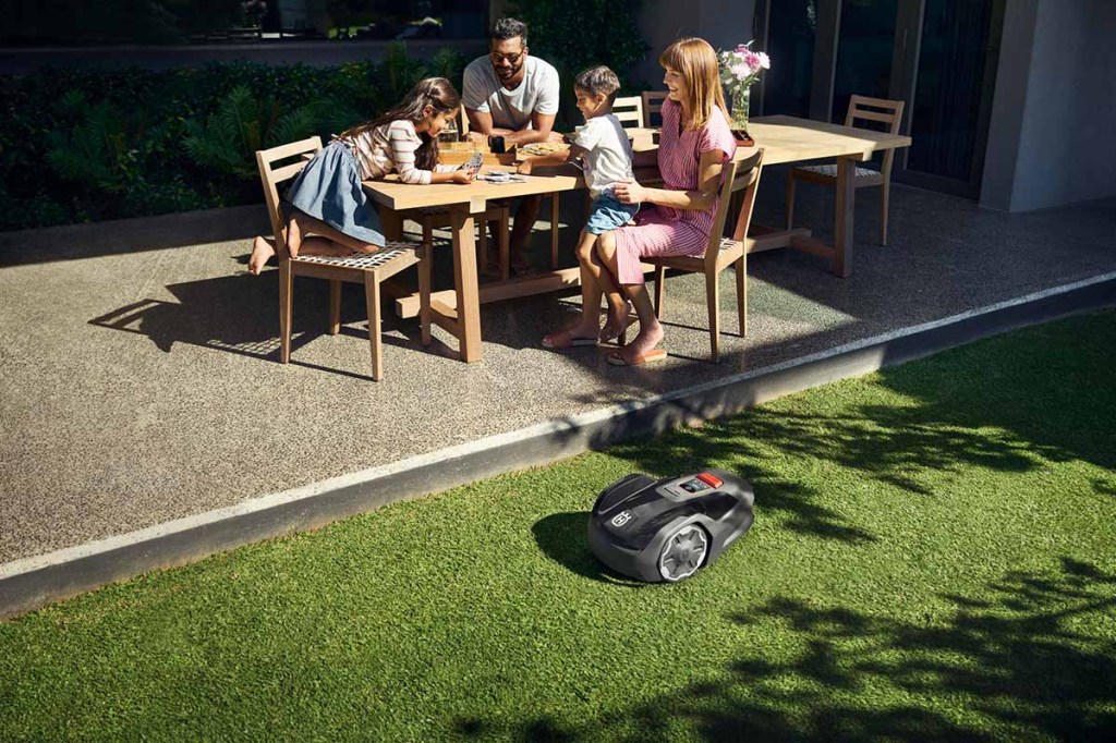 Ein Rasenmäh-Roboter fährt auf Gras neben einer Familie am Tisch.