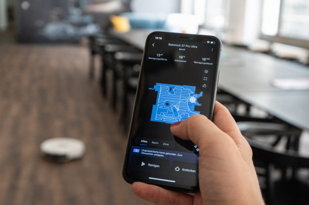 Hand hält dunkles Smartphone mit Raumkarte, im Hintergrund Esstisch und weißer Saug- und Wischroboter auf dunklem Holzboden