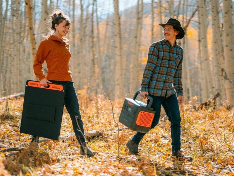 Mann und Frau gehen durch Wald voller Laub mit Solar Panel und Power Station in der Hand