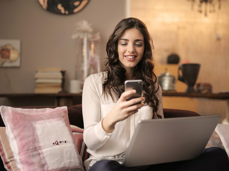 Eine Frau hat einen Laptop auf dem Schoß und schaut lächelnd auf ein Smartphone.