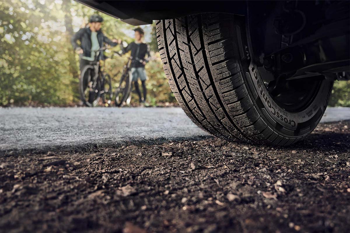 Autoreifen auf einer Schotterpiste von unten im Anschnitt, im hintergrund sieht man zwei Kinder auf einem Fahrrad