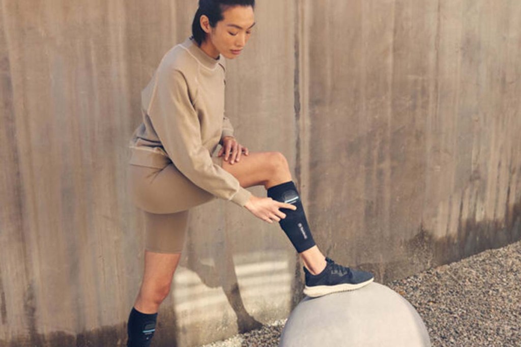 Eine Frau trägt die RecoveryPulse-Beinmanschetten von Therabody. Abgestützt auf eine Kugel schaltet sie die rechte ein.