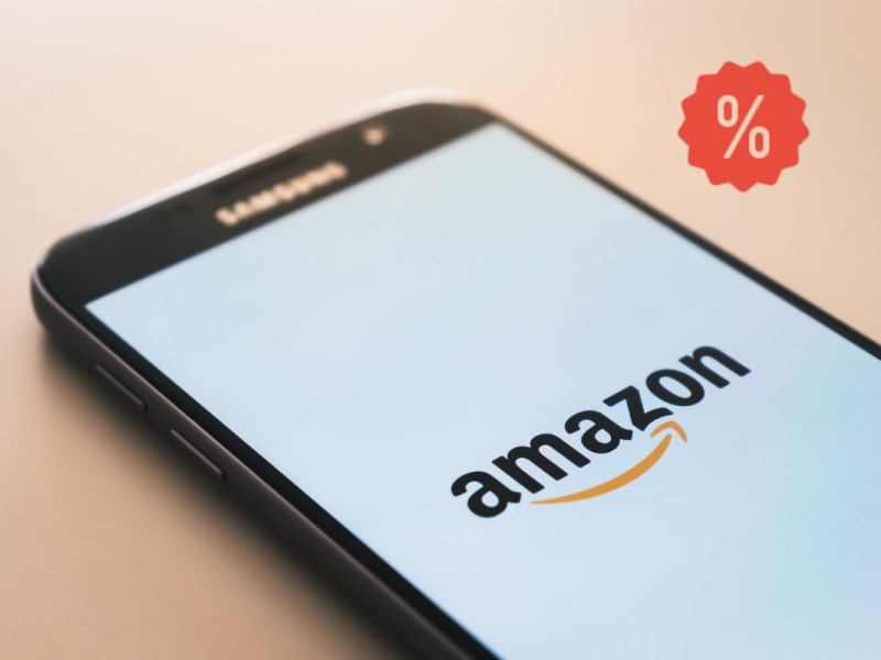 Dunkles Smartphone ragt in Bild rein mit Amazon Logo auf Bildschirm auf hellem Hintergrund mit rotem Prozentzeichen rechts oben