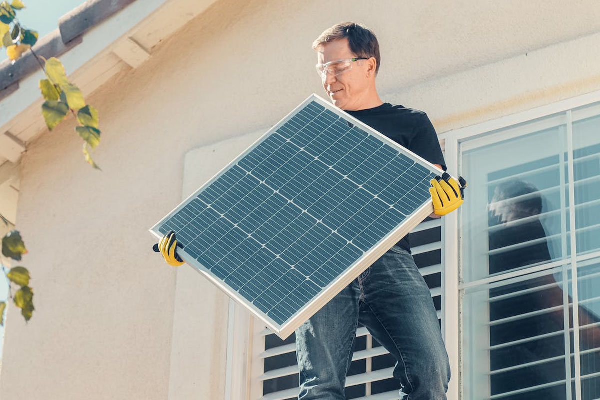 Ein Mann auf einem Hausdach in der Sonne mit einem Solarpanel in der Hand.