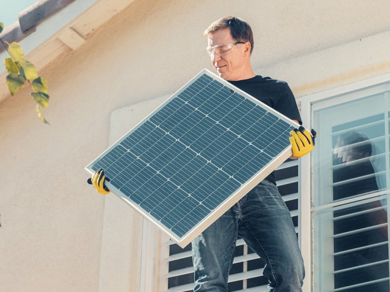 Ein Mann auf einem Hausdach in der Sonne mit einem Solarpanel in der Hand.