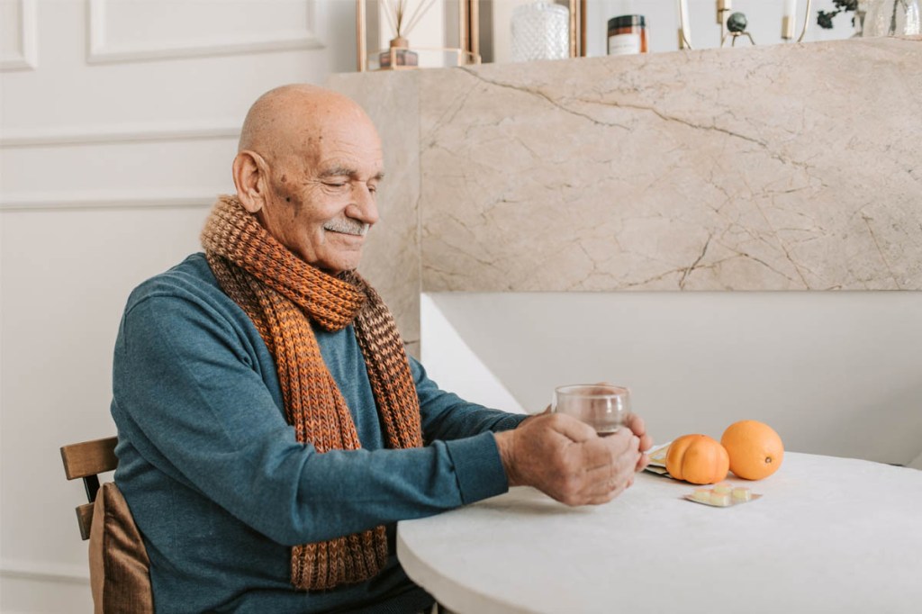 Ein alter Mann mit Schal umfasst genießerisch eine Tasse Tee. Auf dem Tisch liegen zwei Mandarinen und ein paar Tabletten.