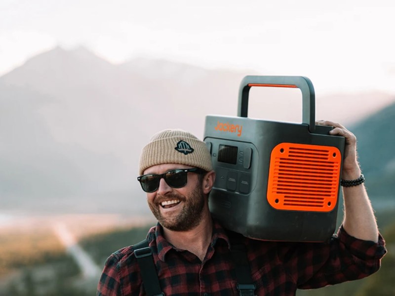 Mann mit Sonnenbrille und Mütze trägt Jackery Power Station in schwarz orange auf Schulter vor Bergkulisse