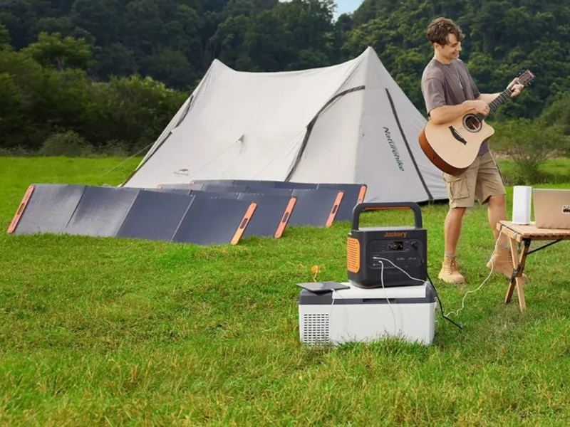 Junger Mann mit Akustikgitarre zwischen Holztisch mit Laptop und orange schwarze Jackery Power Station auf grüner Wiese, im Hintergrund aufgestellte Solarpanels vor weißem Zelt