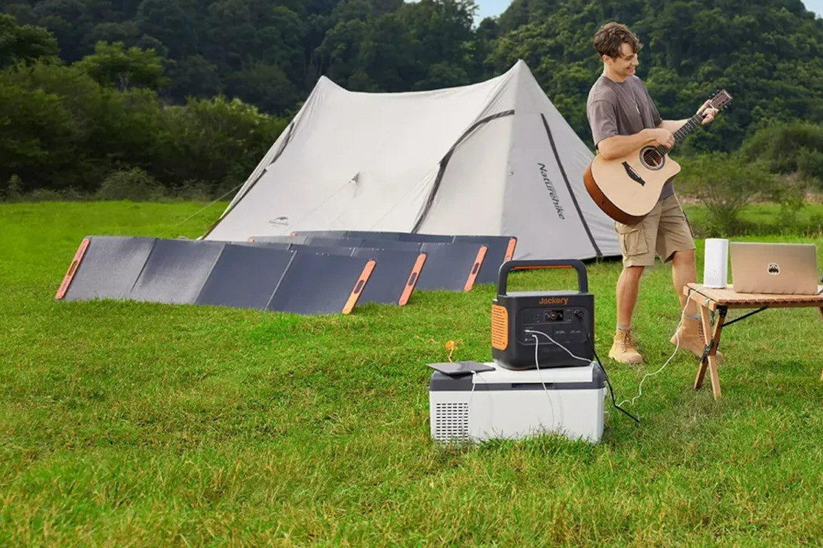 Junger Mann mit Akustikgitarre zwischen Holztisch mit Laptop und orange schwarze Jackery Power Station auf grüner Wiese, im Hintergrund aufgestellte Solarpanels vor weißem Zelt