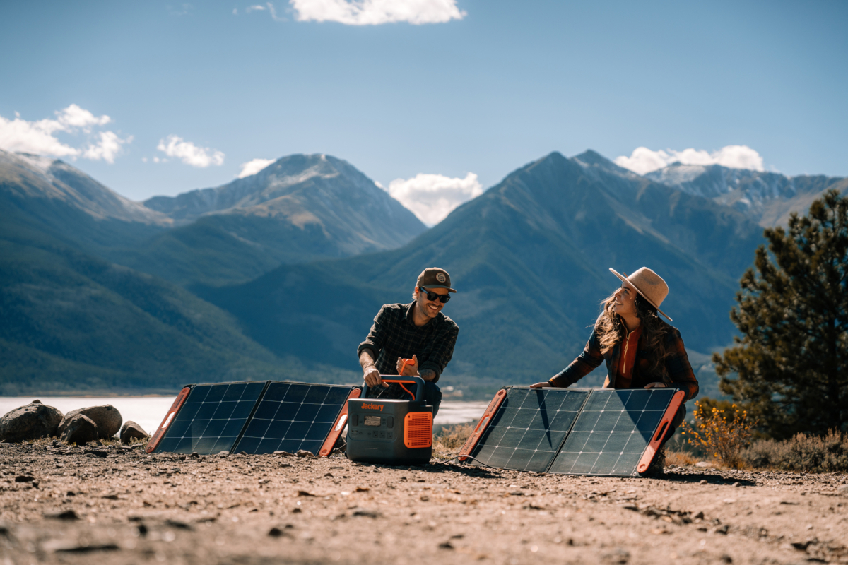 Junge Frau und junger Mann vor Bergpanorama auf Sandboden mit Jackery Power Stationen und zwei Solarpanels