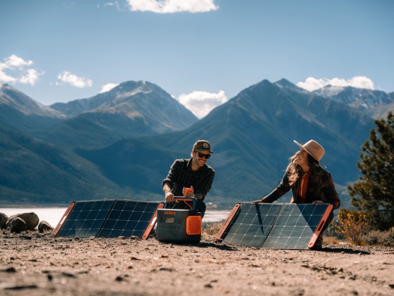 Junge Frau und junger Mann vor Bergpanorama auf Sandboden mit Jackery Power Stationen und zwei Solarpanels