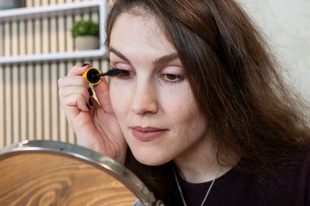 Frau mit langen braunen Haaren vor Schminkspiegel trägt Mascara auf.