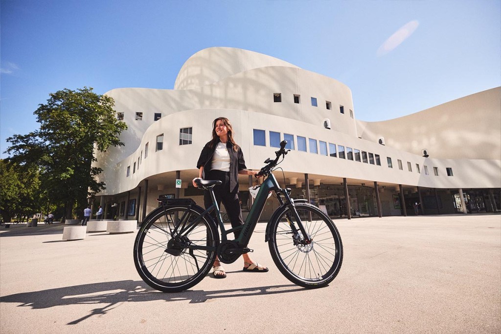 Frau steht hinter ihrem E-Bike, dahinter ein großes Gebäude