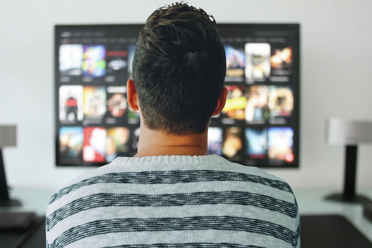 Mann in weiß grau gestreiftem Pullover sitzt mit dem Rücken zur Kamera vor dem großen Fernseher auf dem Film Kacheln zu sehen sind