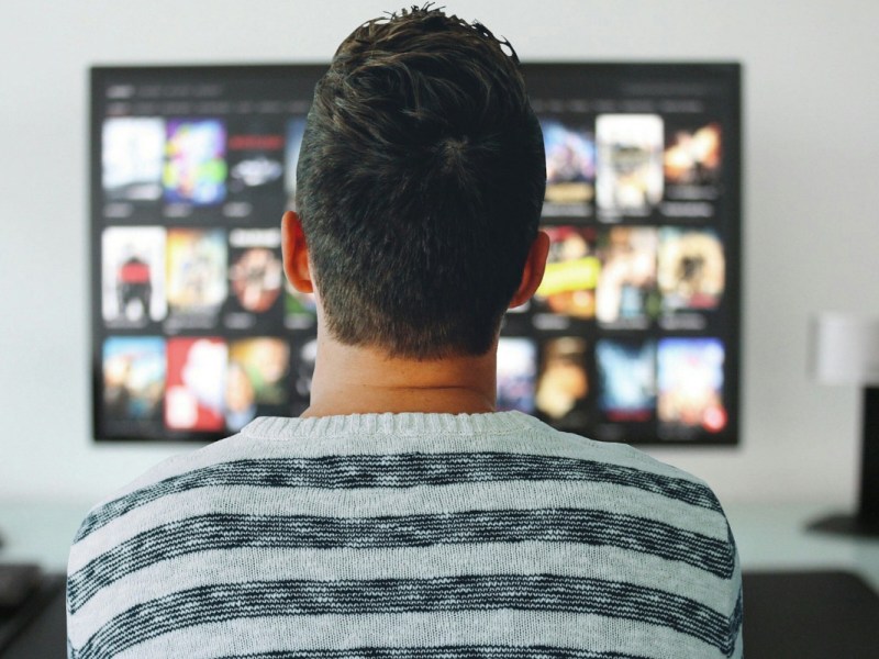Mann in weiß grau gestreiftem Pullover sitzt mit dem Rücken zur Kamera vor dem großen Fernseher auf dem Film Kacheln zu sehen sind