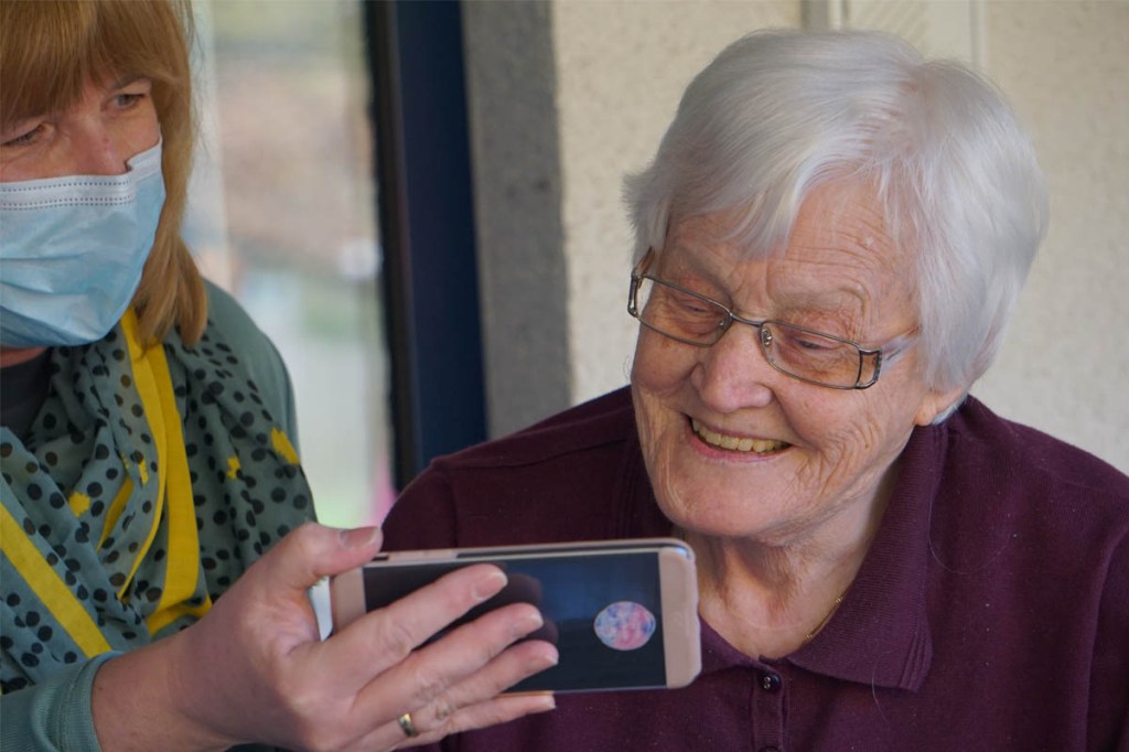 Eine alte Frau schaut lachend auf ein Handy, das eine jüngere ihr hinhält.