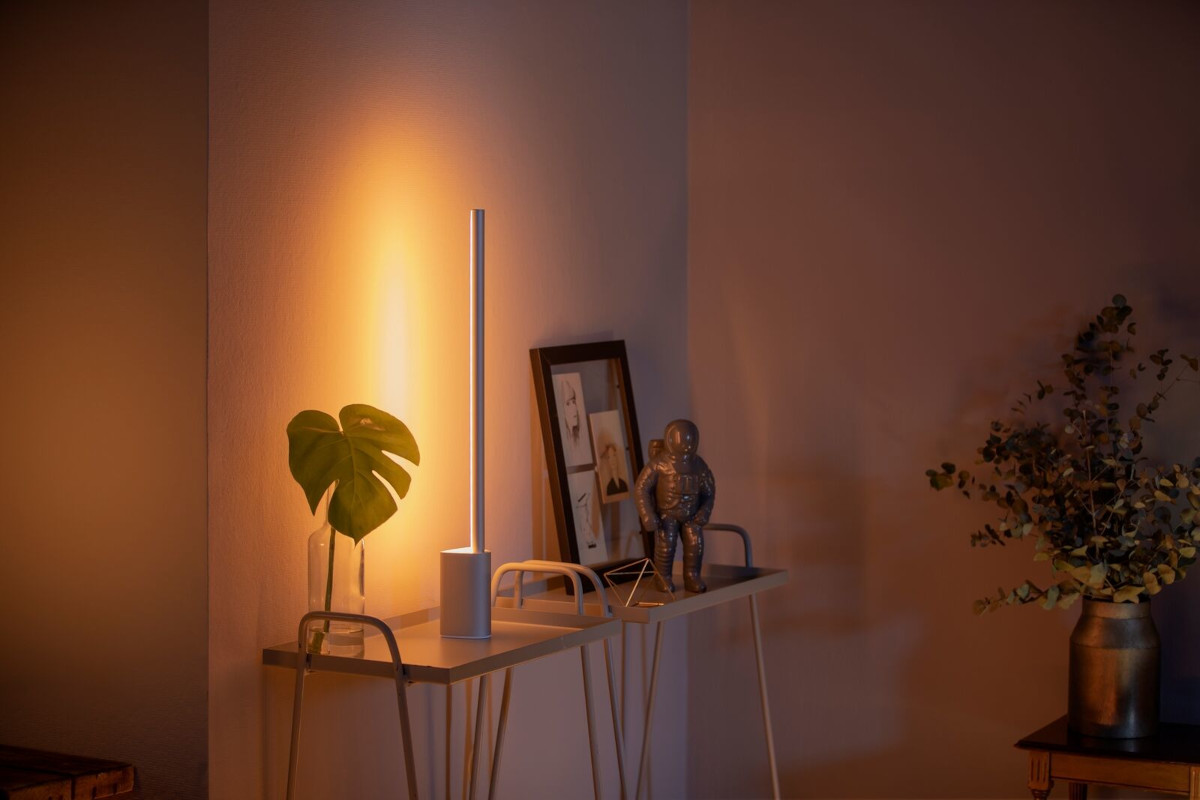 Weiße längliche Philips-Hue-Tischleuchte auf Kommode an der Wand neben Pflanze und Bilderrahmen leuchtet orange