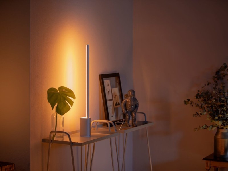 Weiße längliche Philips-Hue-Tischleuchte auf Kommode an der Wand neben Pflanze und Bilderrahmen leuchtet orange