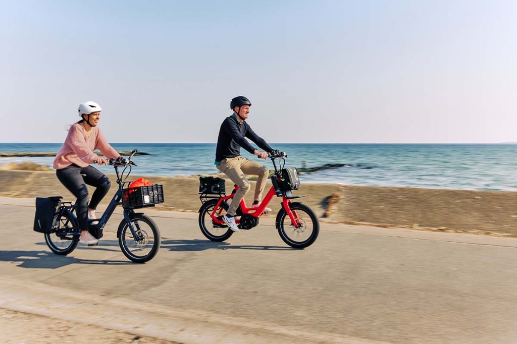 zwei Personen fahren auf Kompakt-E-bike an Strandpromenade lang, im HIntergrund sieht man das Meer