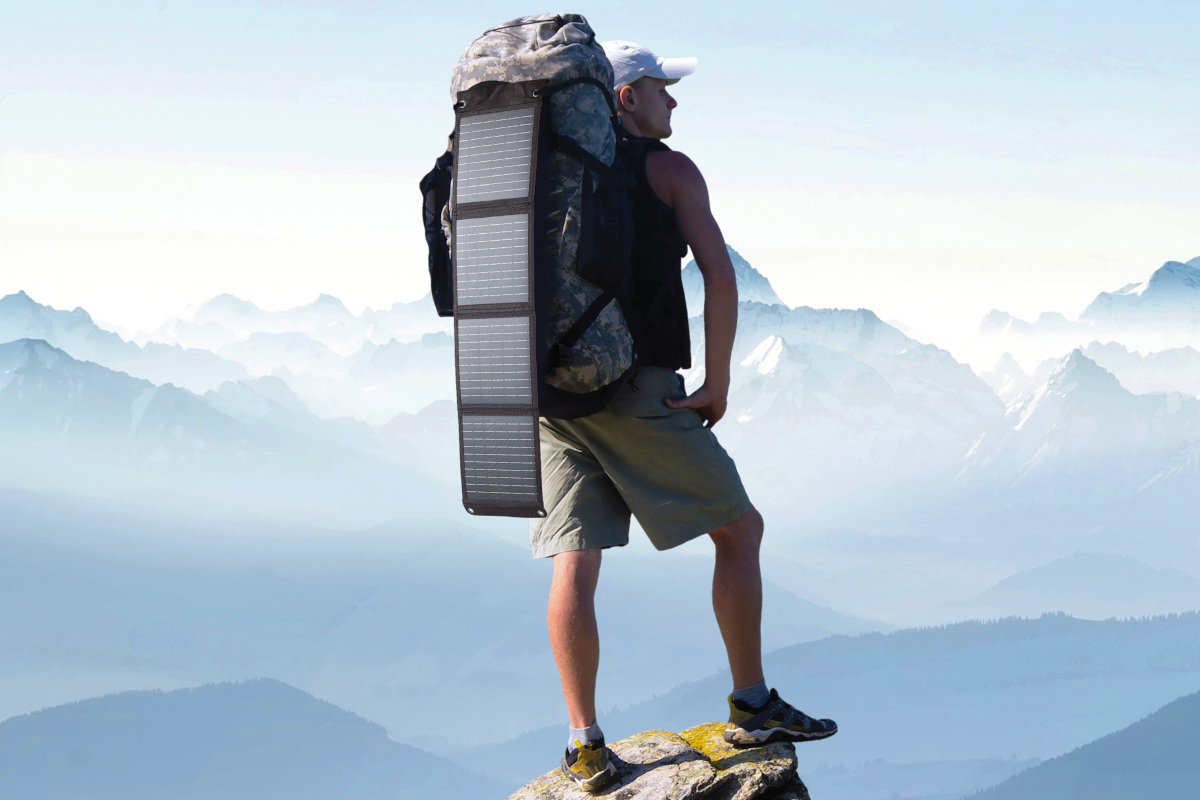 Mann von hinten mit Wanderrucksack an dem ausgeklapptes Solarmodul hängt auf Bergspitze vor blauem Bergpanorama