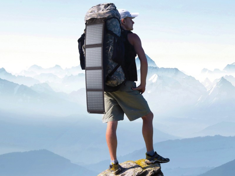 Mann von hinten mit Wanderrucksack an dem ausgeklapptes Solarmodul hängt auf Bergspitze vor blauem Bergpanorama