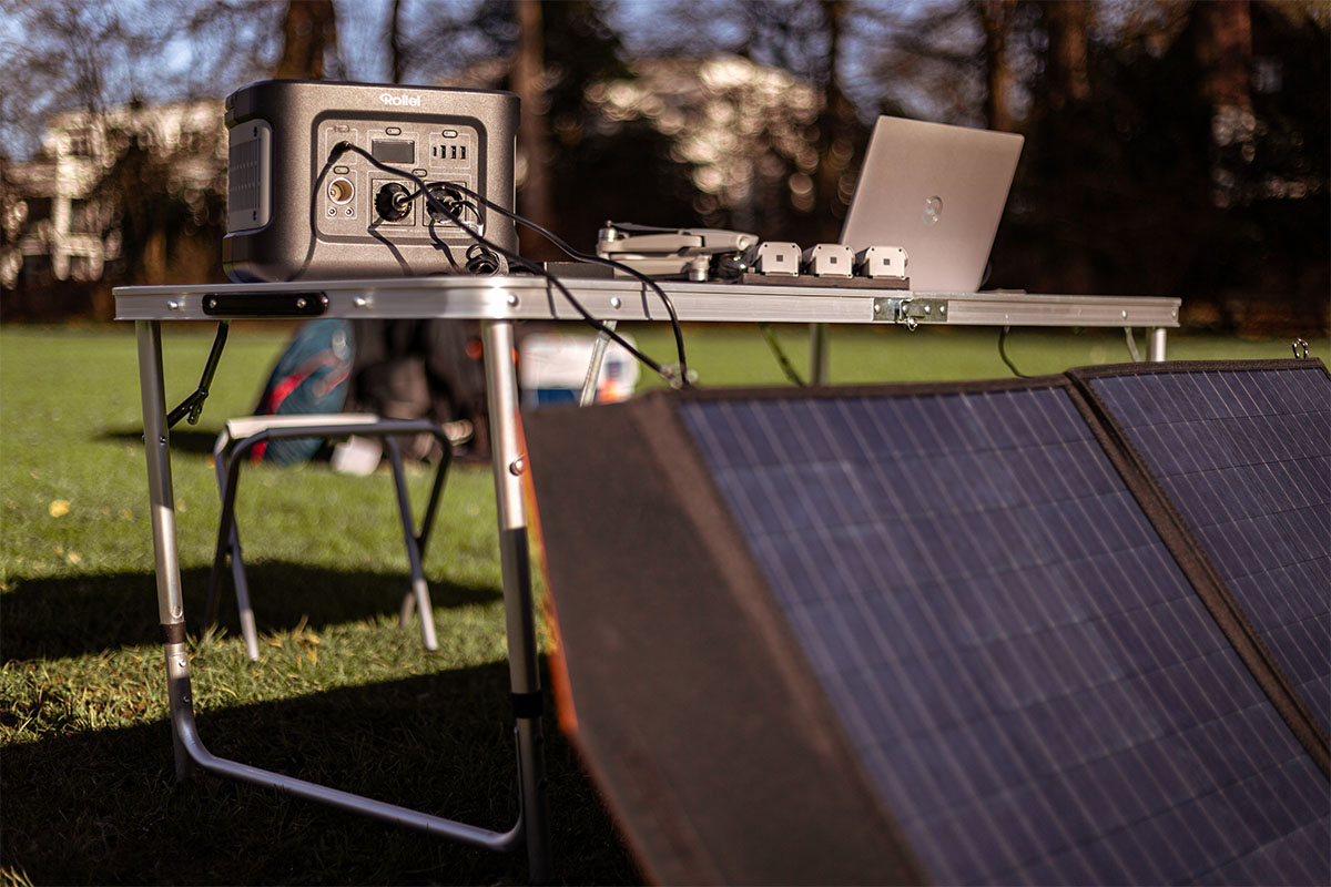Auf einer Wiese steht ein Tisch mit Elektrogeräten und einer Rollei Power Station. Davor ist ein Solarpanel platziert.