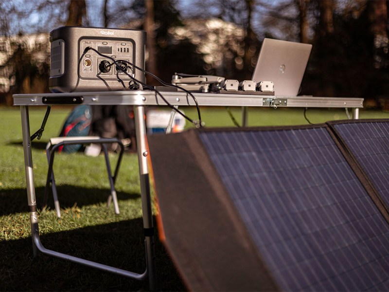 Auf einer Wiese steht ein Tisch mit Elektrogeräten und einer Rollei Power Station. Davor ist ein Solarpanel platziert.