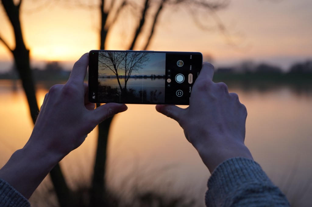Zwei Hände halten Smartphone waagerecht um Foto von Sonnenuntergang am See zu machen