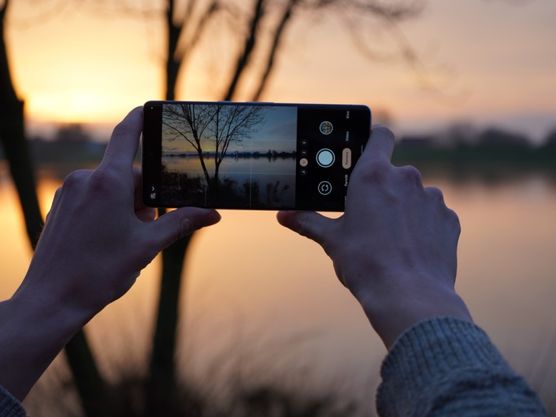 Zwei Hände halten Smartphone waagerecht um Foto von Sonnenuntergang am See zu machen