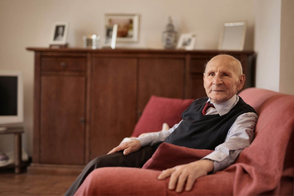 Ein alter Mann sitzt auf einem roten Sofa, den Arm auf die Lehne gestützt.