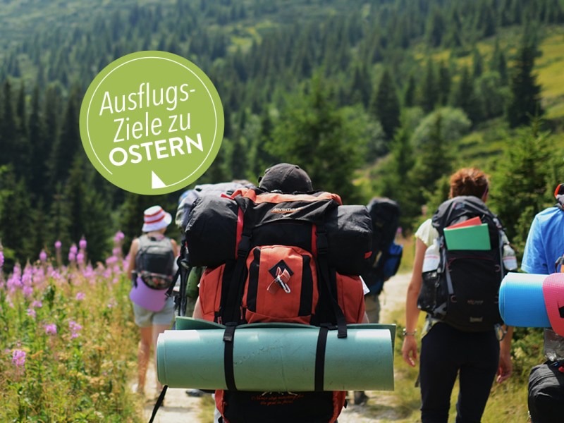 Gruppe von Menschen mit Rucksäcken wandern durch Berglandschaft; grüner Stempel "Ausflugsziele zu Ostern"