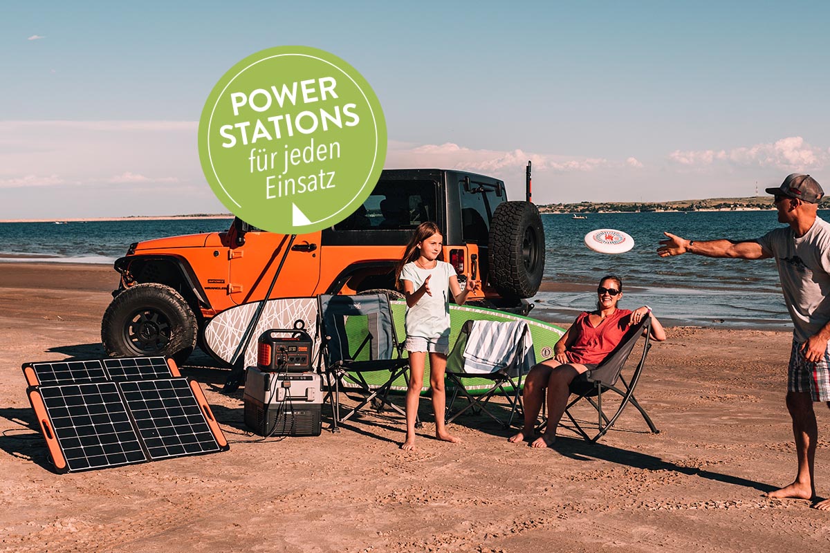 Familie spielt Frisbee am Strand vor orangenem Geländewagen, aufgestellten Solarpanels, Powerstation und Surfbrett