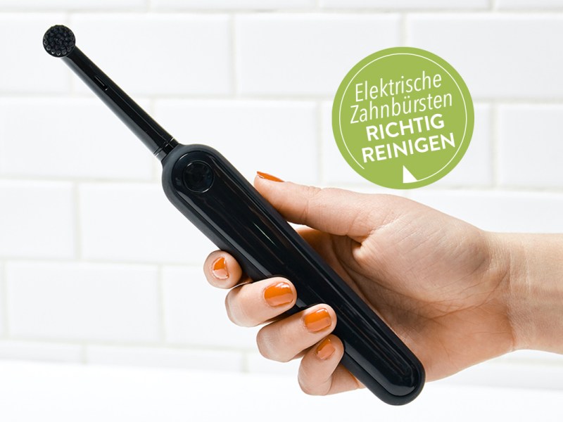 Hand mit roten Fingernägeln hält schwarze elektrische Zahnbürste vor weißer Steinwand; grüner Button oben rechts "Elektrische Zahnbürste richtig reinigen"