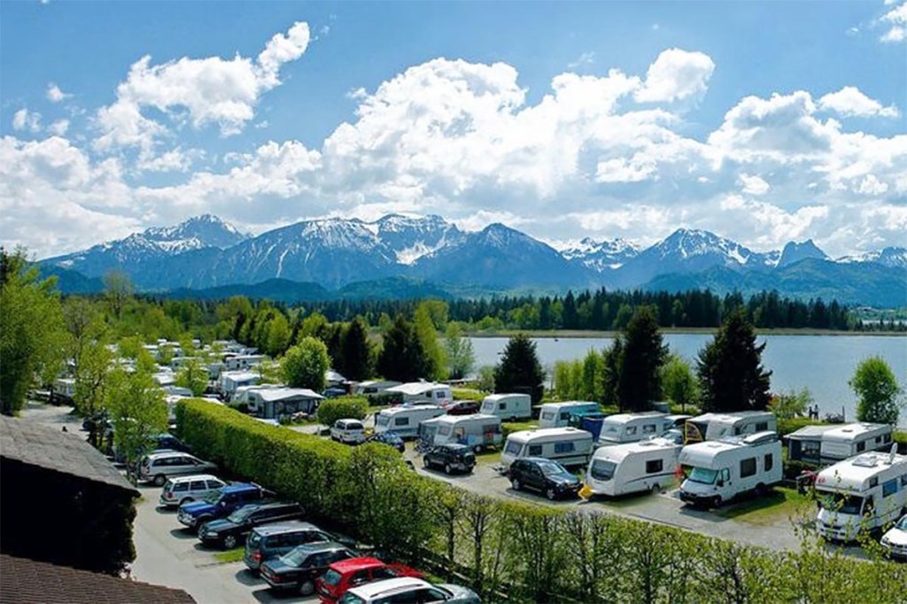 Parkende Auots und Wohnwagen zwischen Bäumen und Hecken vor See und großen Bergen im Hintergrund