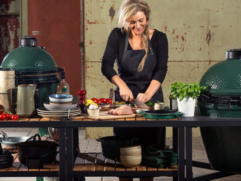 Blonde Frau mit schwarzer Schürze an Kochtisch bereitet Lebensmittel zu. Links und rechts zwei große ovale grüne Keramikgrills