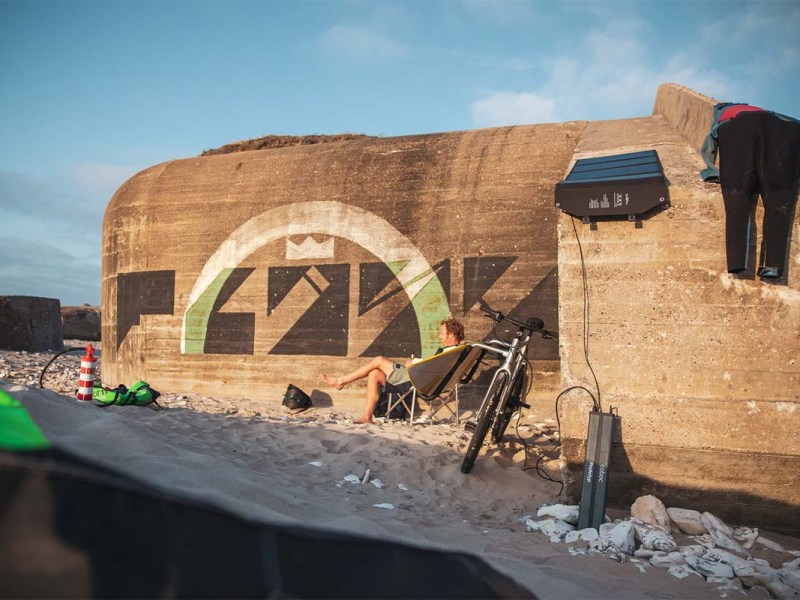 E-Bike Fahrer sitzt am Strand vor einer Mauer, neben im sein E-Bike, welches von einem Akku geladen wird