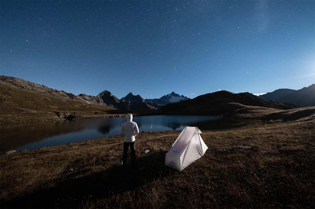 Mann steht nachts mitten in der Natur, neben ihm sein weißes Zelt, er blickt in die Ferne