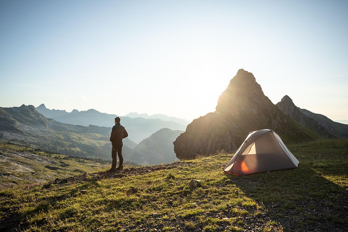 Mann steht vor Zelt in Gebirgslandschaft.