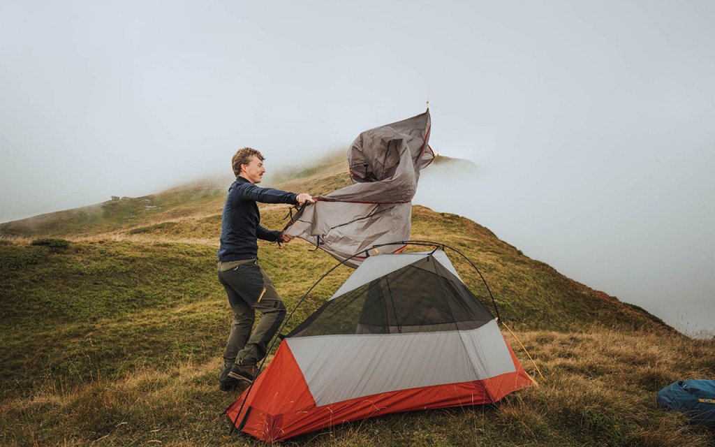 Mann baut Zelt in Gebirgslandschaft bei starkem Wind und Nebel auf.