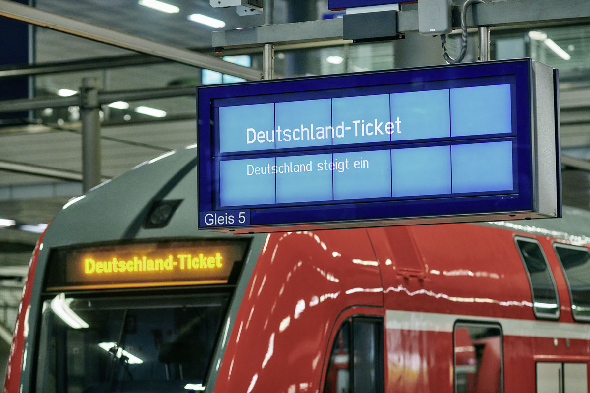 Informationsanzeige an einem Bahnsteig mit Regionalzug im Hintergrund.