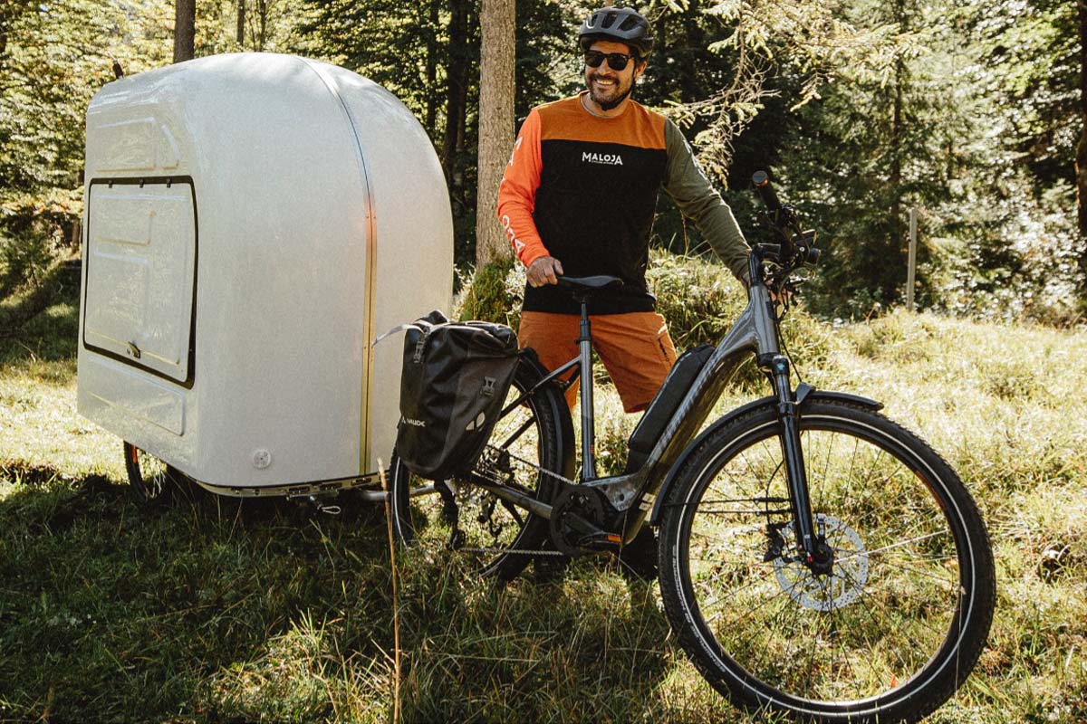 Mann steht neben seinem E-Bike im Wald, hinter seinem Rad ist ein kleiner Wohnmobil-Anhänger, der vom Rad gezogen wird