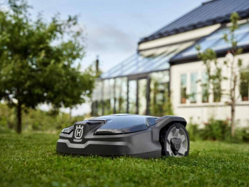 Dunkler Mähroboter auf Rasen von vorne vor hellem Haus und grünem Baum
