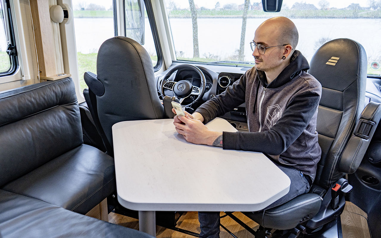 Mann sitz in einem Wohnmobil am Esstisch und schaut auf ein Smartphone.