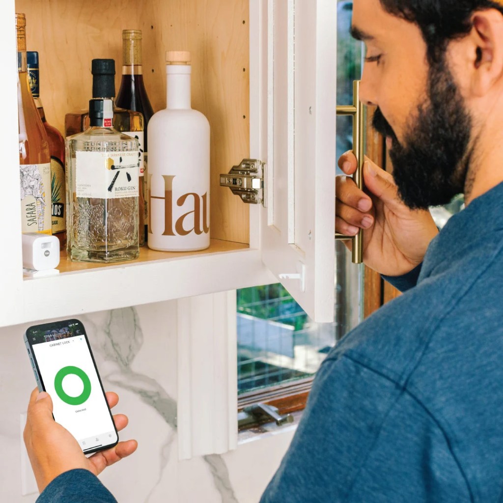 Mann mit Smartphone und aktiver Yale-App vor einem halb geöffneten Schrank mit Spiritousen