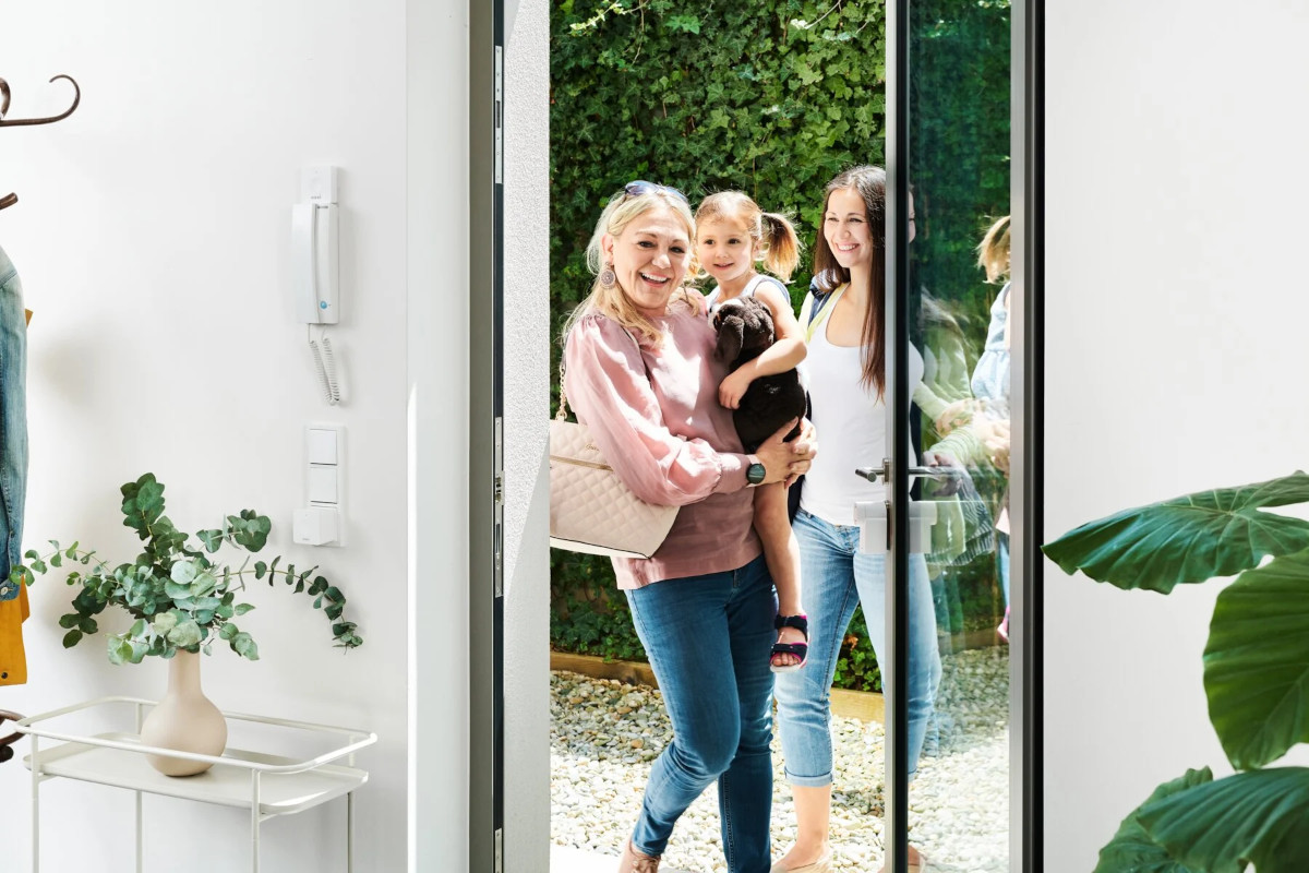 Lächelnde Frau mit Mädchen auf Arm vor zweiter Frau stehen vor geöffneter Glastür an der ein weißes Nuki-Türschloss angebracht ist