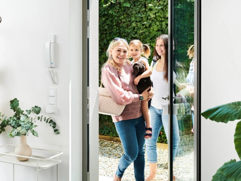 Lächelnde Frau mit Mädchen auf Arm vor zweiter Frau stehen vor geöffneter Glastür an der ein weißes Nuki-Türschloss angebracht ist
