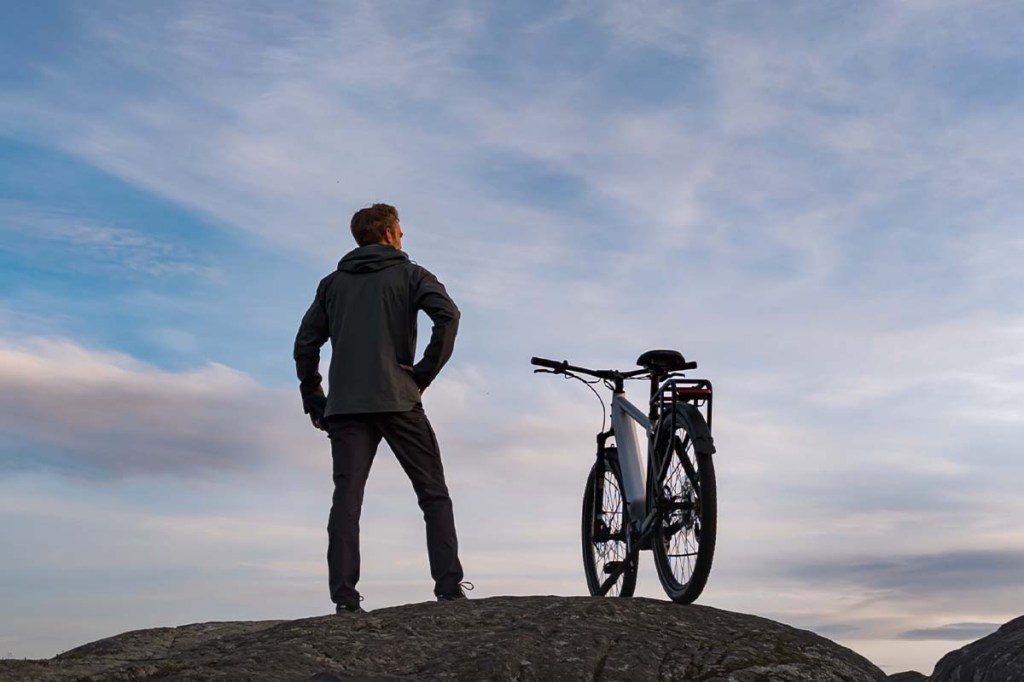 Mann neben seinem E-bike auf Bergkuppe stehend, guckt in die Ferne