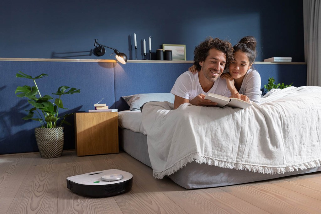 Ein Paar liegt lesend auf einem Bett. Der Vorwerk Kobold VR7 fährt auf dem Boden des Schlafzimmers.
